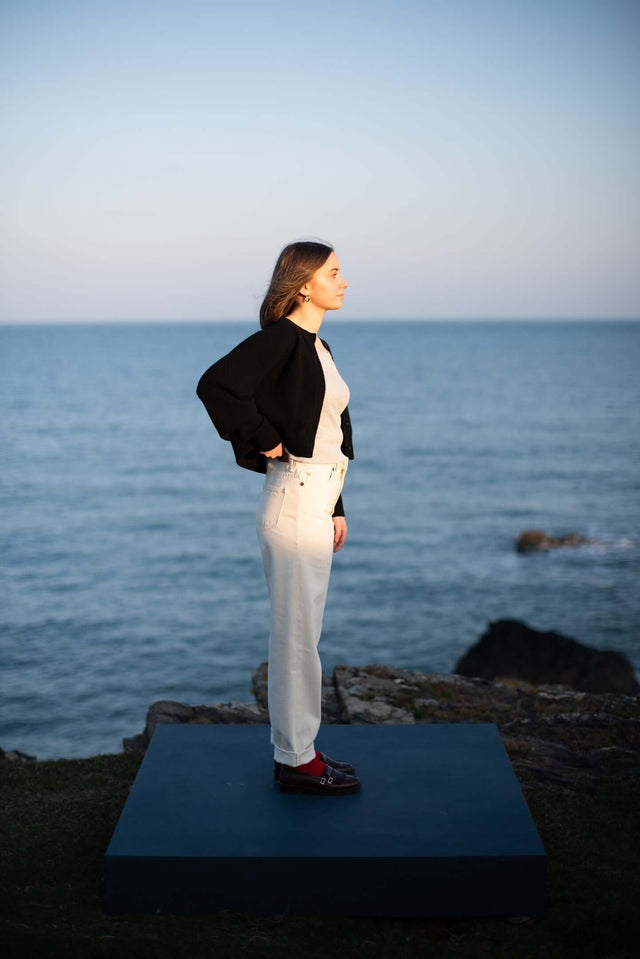 Person standing on a blue mat by the ocean with a black jacket and white pants.