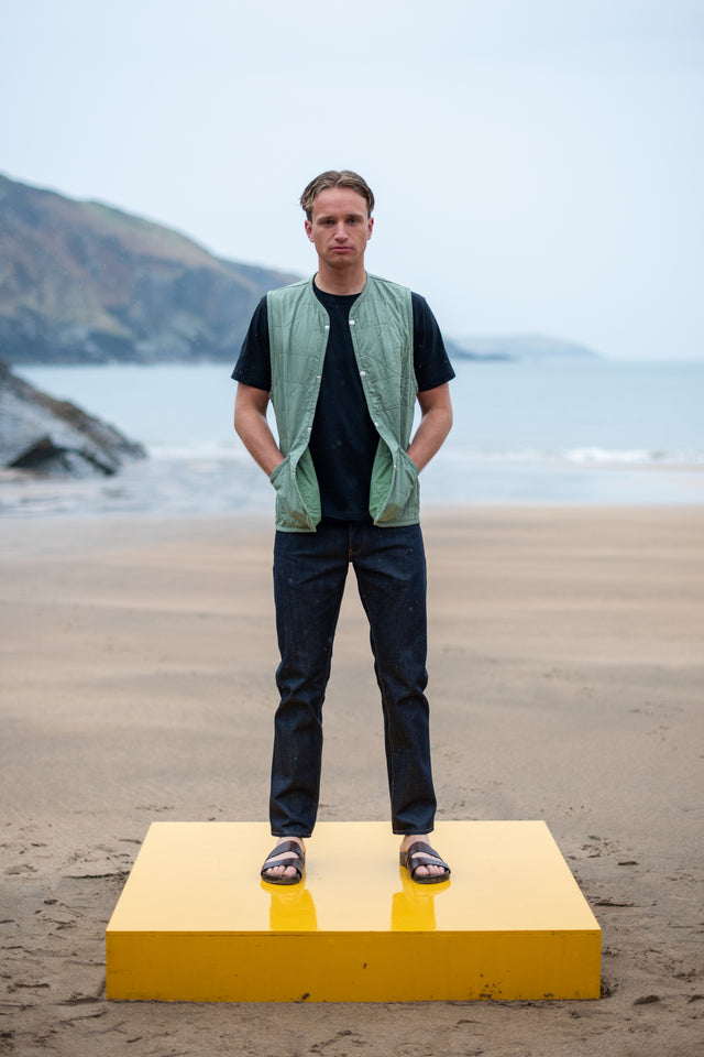 Man standing on a yellow platform at the beach wearing a green vest and black shirt.
